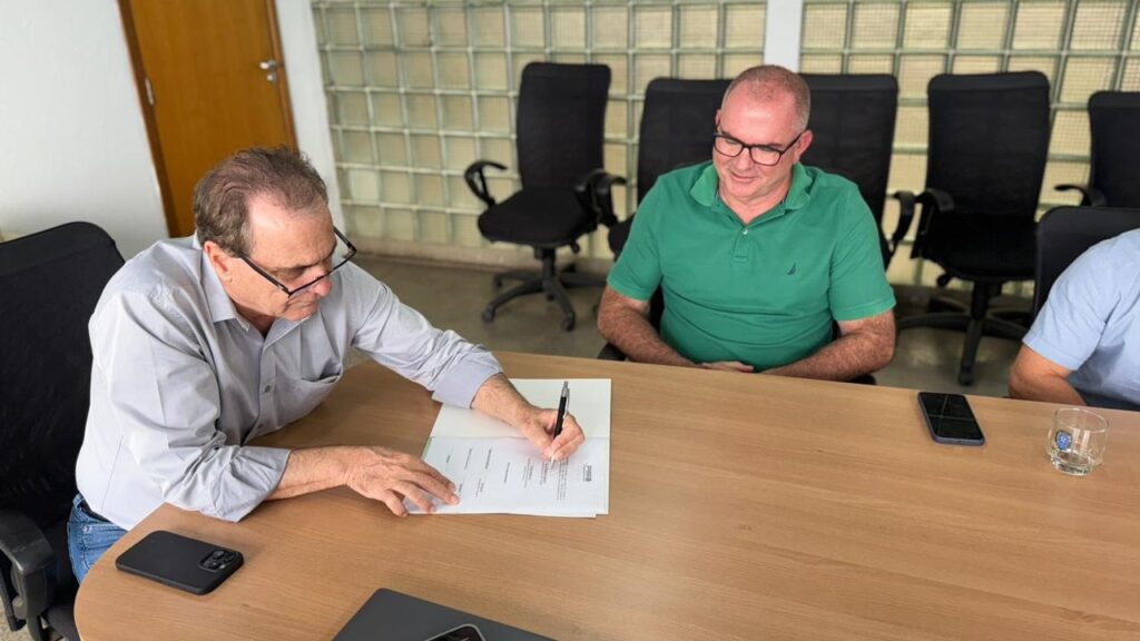Na imagem, dois homens estão sentados à mesa em uma sala de reuniões. Um deles, vestindo uma camisa cinza e óculos, está assinando um documento. O outro homem, vestindo uma camisa verde e óculos, sorri enquanto observa. Há cadeiras de escritório pretas ao fundo e uma porta de madeira à esquerda. Um celular e um copo com um líquido transparente estão sobre a mesa.
