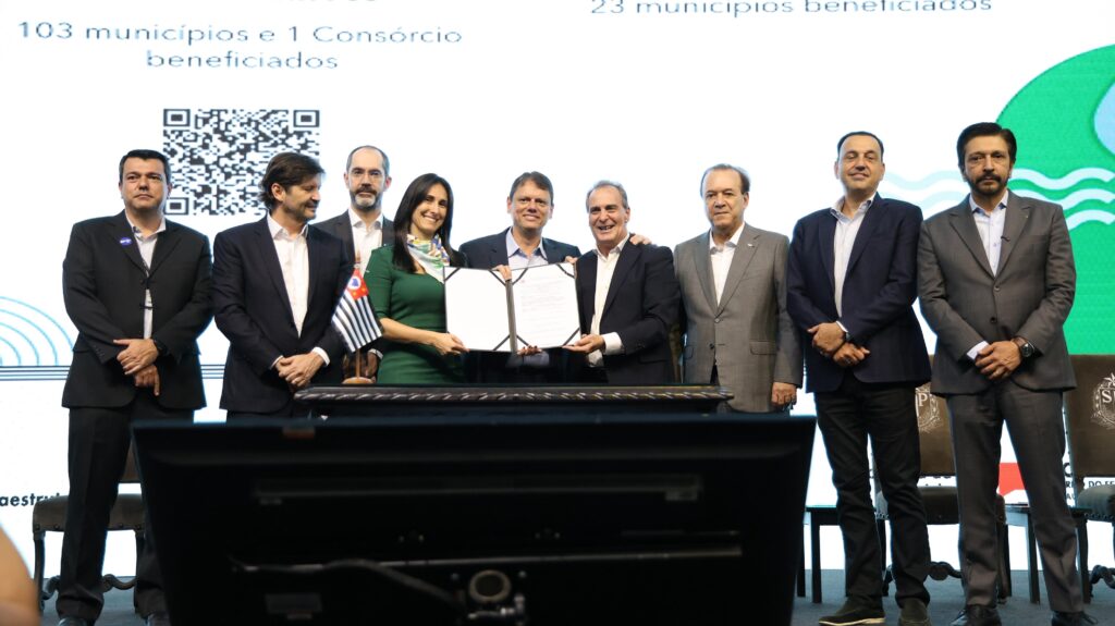 A imagem mostra um grupo de pessoas, a maioria homens vestidos com ternos, em pé atrás de uma mesa onde um documento está sendo assinado. Uma mulher em um vestido verde está no centro, segurando o documento junto com um homem. Há bandeiras e um QR code visível ao fundo, sugerindo um evento oficial ou uma cerimônia de assinatura de acordo. O texto ao fundo parece indicar que 103 municípios e 1 consórcio foram beneficiados.