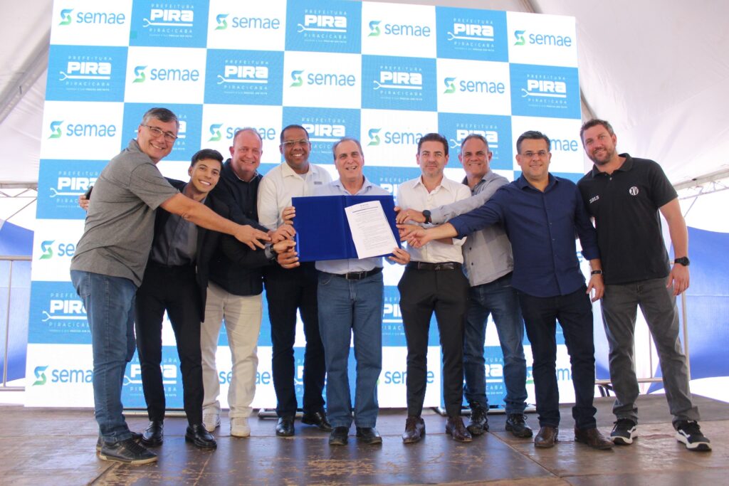 A imagem mostra um grupo de dez homens em pé, todos sorrindo e olhando para a câmera em frente a um painel com o logotipo "PREFEITURA PIRA PIRACICABA" e "semae". Eles estão segurando um documento azul aberto com as mãos entrelaçadas. A maioria dos homens está vestindo camisas de botões e calças, enquanto alguns usam camisetas. O homem no centro, segurando o documento, está com uma camisa azul e jeans. O homem à direita dele usa uma camiseta preta com o número "1913" e um logotipo. O painel de fundo é composto por quadrados azuis e brancos com o logotipo repetido. A iluminação sugere que o evento está acontecendo ao ar livre, possivelmente durante o dia.