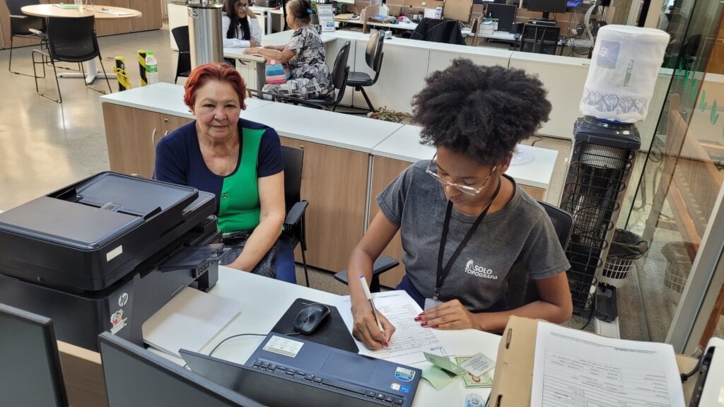 A imagem mostra duas mulheres sentadas em uma mesa em um escritório. A mulher mais velha, à esquerda, tem cabelo ruivo e usa uma blusa azul e verde. Ela está sentada em frente a uma impressora preta. A mulher mais jovem, à direita, tem cabelo cacheado escuro e usa óculos e uma camiseta cinza com um logotipo que diz "SOLO TOPOGRAFIA". Ela está escrevendo em um formulário enquanto uma pilha de documentos e cartões verdes está ao lado dela. Há também um laptop aberto na mesa. Ao fundo, outras pessoas estão trabalhando em seus computadores em um ambiente de escritório. Há também um dispensador de água com um galão de água em cima, à direita da mulher mais jovem.