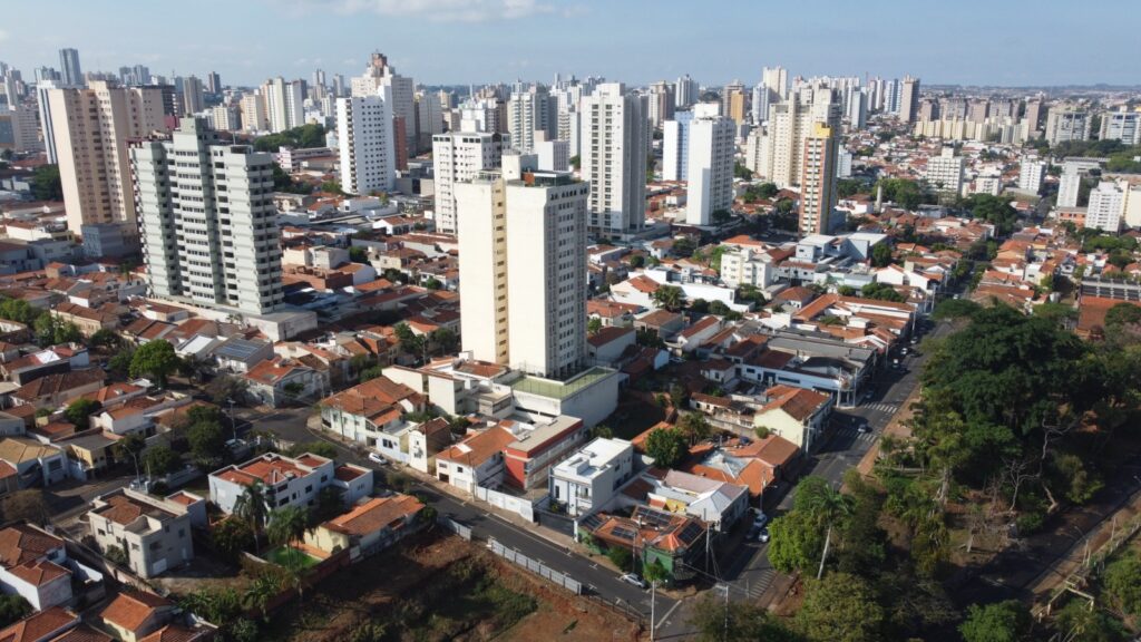 A imagem exibe uma vista aérea de uma paisagem urbana densamente povoada, caracterizada por uma mistura de edifícios residenciais de baixa e média altura com telhados de barro, e arranha-céus modernos que se erguem ao fundo. A arquitetura varia, com casas apresentando fachadas brancas e telhados vermelhos, contrastando com os edifícios de escritórios e apartamentos com estruturas mais contemporâneas, muitas delas em tons de branco e cinza. A vegetação é visível em alguns pontos, com árvores frondosas e áreas verdes pontuando a paisagem urbana, especialmente em primeiro plano e em alguns espaços entre os edifícios. A presença de ruas e a disposição dos edifícios sugerem um planejamento urbano organizado, com uma clara separação entre áreas residenciais e comerciais de maior porte. O céu azul com algumas nuvens leves completa a cena, indicando um dia ensolarado.