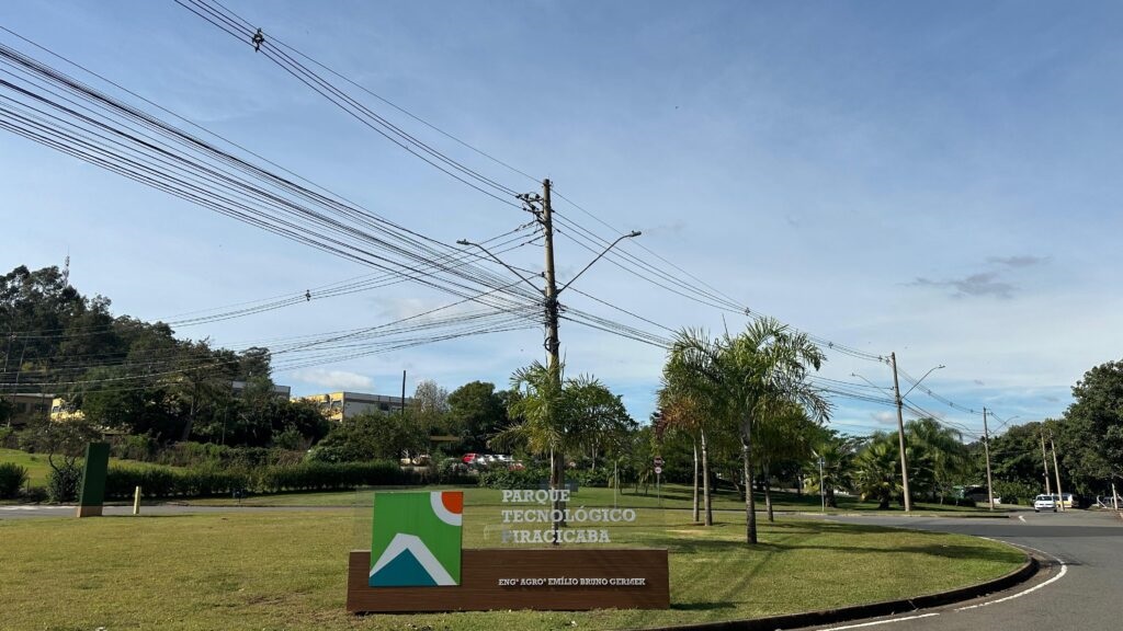 A imagem mostra a entrada de um parque tecnológico. Há um letreiro com o nome "PARQUE TECNOLÓGICO PIRACICABA" e, abaixo, "ENG. AGRO EMÍLIO BRUNO GERMER". O letreiro tem um design moderno com formas geométricas em verde, laranja e azul, sobre uma base de madeira. O local é arborizado, com gramados bem cuidados e palmeiras. Ao fundo, observam-se edifícios e uma estrada com alguns carros. Uma grande quantidade de fios elétricos cruza o céu, passando por postes de iluminação, o que sugere a infraestrutura de um centro moderno. O céu está parcialmente nublado, com tons de azul claro.