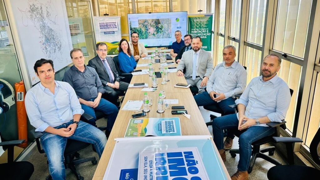 A imagem mostra um grupo de homens e uma mulher sentados ao redor de uma longa mesa de madeira em uma sala de reuniões. Há garrafas de água e alguns papéis sobre a mesa. Ao fundo, há um mapa em uma parede e um telão exibindo um mapa com alguns dados. Há também um banner com o texto "NÃO COMPRE IMÓVEL IRREGULAR". As pessoas parecem estar em uma reunião ou discussão.