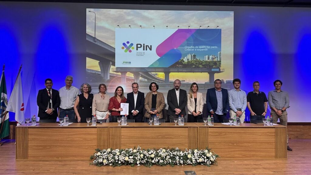 A imagem mostra um grupo de 11 pessoas, 6 mulheres e 5 homens, posando para uma foto atrás de um balcão de madeira. Todos estão de pé, exceto por uma mulher que está sentada em uma cadeira. O grupo parece estar em um evento formal, possivelmente uma cerimônia de premiação ou uma assinatura de acordo, pois há bandeiras à esquerda e um grande banner ao fundo. O banner exibe um logotipo com as letras "PiN" e a frase "Orgulho de quem faz parte. Crescer e expandir.". As pessoas estão vestidas de forma profissional, com ternos, blazers e vestidos. Há garrafas de água e copos sobre o balcão. O ambiente é iluminado e o fundo é um palco com cortinas azuis.