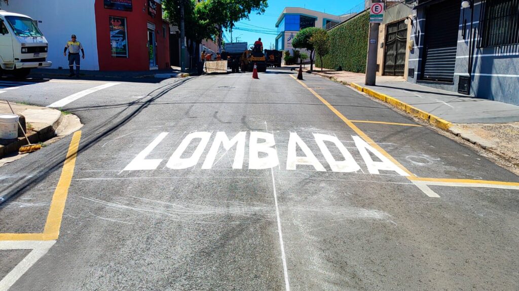 A imagem mostra uma rua em um dia ensolarado, com marcações de trânsito sendo feitas ou recém-feitas. No centro da imagem, em letras brancas grandes e em caixa alta, está pintada a palavra "LOMBADA", indicando a presença de uma lombada (ou redutor de velocidade) na via. Há linhas brancas e amarelas pintadas no asfalto, sugerindo que a sinalização de trânsito está sendo atualizada ou mantida. Ao fundo, observam-se alguns trabalhadores e máquinas, como um rolo compactador, indicando que o trabalho de pavimentação ou sinalização está em andamento. Edifícios comerciais e residenciais ladeiam a rua, com uma fachada vermelha à esquerda e uma com detalhes em azul e verde à direita. Um veículo branco e um caminhão também são visíveis na cena, além de cones de sinalização. A calçada é visível em algumas partes, com um poste e uma placa de trânsito (com a letra "E" dentro de um círculo vermelho) à direita. Um balde branco e uma vassoura estão posicionados perto da calçada à esquerda.