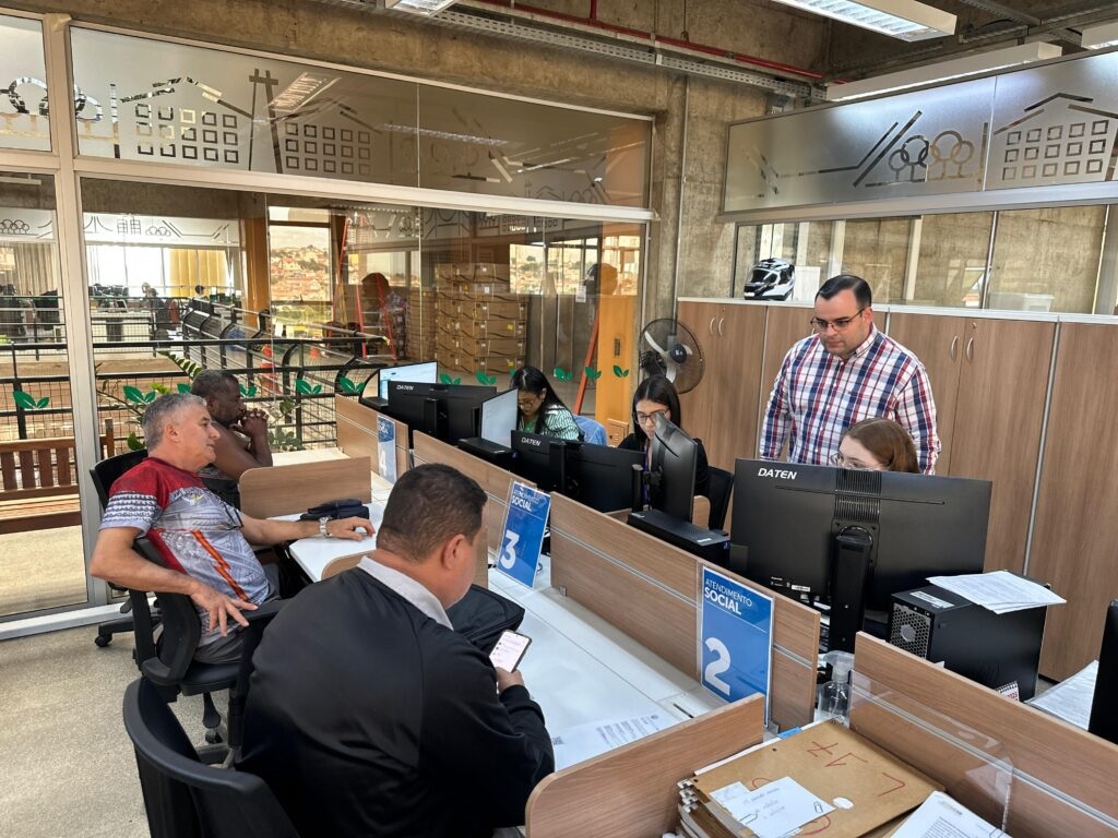 A imagem mostra um escritório com várias pessoas trabalhando em computadores. Há divisórias de mesa, e os funcionários parecem estar em um ambiente de atendimento ao público, com sinalização indicando "ATENDIMENTO SOCIAL". As paredes são de concreto aparente e há janelas grandes que permitem a entrada de luz natural, com vista para uma paisagem externa. Em primeiro plano, à esquerda, um homem de camisa colorida está sentado e olhando para o lado. Ao seu lado, um homem negro também está sentado, com as mãos no queixo. Mais à direita, um homem de terno escuro está olhando para o celular. No centro da imagem, um homem com óculos e camisa xadrez azul e vermelha está em pé, observando o trabalho de duas mulheres sentadas em frente aos computadores. Uma delas, à esquerda, tem óculos e veste uma blusa verde, enquanto a outra, à direita, tem cabelo ruivo e veste uma blusa branca. As mesas possuem monitores, teclados e papéis. Ao fundo, é possível ver mais mesas de trabalho e prateleiras com caixas. A decoração do escritório inclui alguns adesivos nas janelas com desenhos de casas e árvores. Um ventilador de mesa está posicionado em uma das áreas de trabalho.