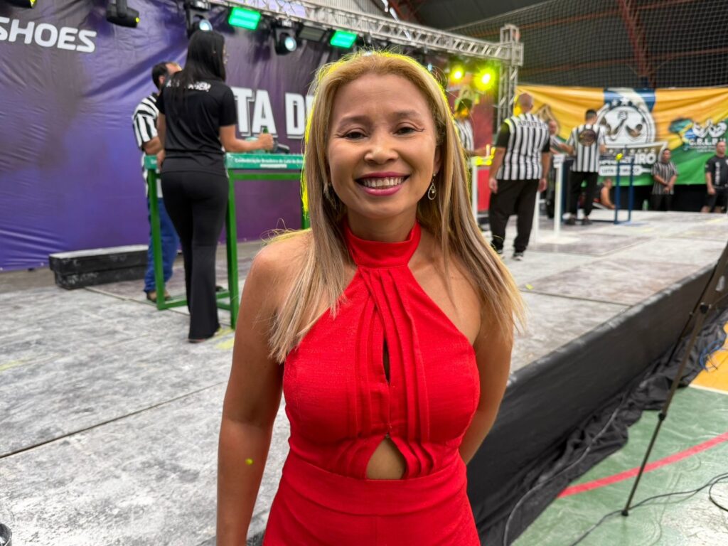 A mulher sorri para a câmera em um close-up, vestindo um vestido vermelho vibrante com um decote halter e recortes na barriga. Seu cabelo loiro escorrega pelos ombros e ela usa brincos pendentes. O fundo está um pouco desfocado, mas mostra uma cena de evento com pessoas vestindo uniformes de árbitro listrados em preto e branco, mesas de competição e um banner com o logotipo de um evento de luta de braço. As luzes do palco brilham, criando uma atmosfera animada.