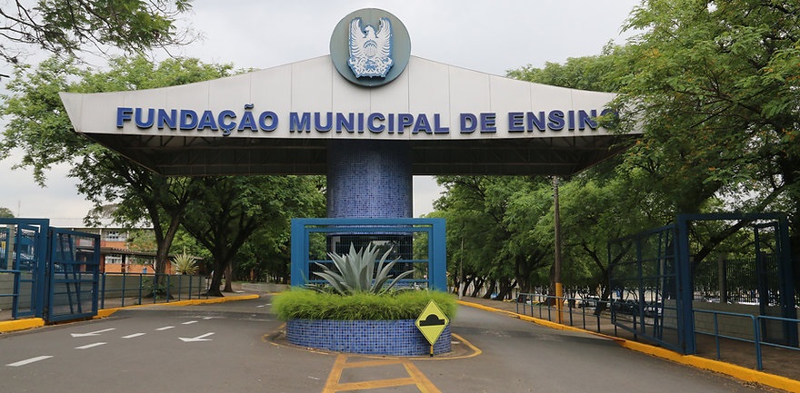 A imagem mostra a entrada principal de uma instituição, identificada por um letreiro luminoso com a inscrição "FUNDAÇÃO MUNICIPAL DE ENSINO". O letreiro está fixado sobre um pórtico com colunas revestidas de azulejos azuis. À frente do pórtico, há um canteiro circular com plantas ornamentais, incluindo uma agave, também cercado por azulejos azuis. A entrada é ladeada por portões de metal azuis e cercas do mesmo material. A via de acesso à fundação é pavimentada e possui sinalização de trânsito, incluindo setas brancas no asfalto e uma placa amarela indicando um redutor de velocidade. Ao fundo, vislumbra-se a vegetação exuberante e parte da edificação da instituição. O céu está nublado, conferindo uma iluminação difusa à cena.