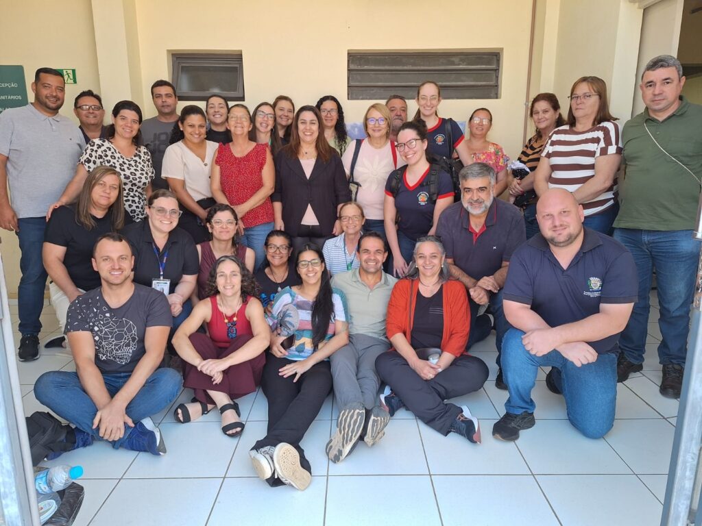 A imagem mostra um grupo de cerca de 30 pessoas, em sua maioria adultos, posando para uma foto. Eles estão em pé e sentados, em um ambiente externo com uma parede de cor clara ao fundo. A maioria das pessoas está vestindo roupas casuais, e algumas parecem ser funcionários de alguma instituição, pois alguns usam crachás ou uniformes. A atmosfera parece ser amigável e descontraída.