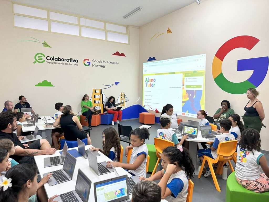 A imagem mostra uma sala de aula com várias crianças e adultos, todos focados em computadores. A sala é decorada com elementos relacionados à tecnologia e educação, incluindo o logo do Google e a frase "Google for Education Partner". No centro da sala, um projetor exibe uma tela com o título "Aluno Tutor", sugerindo uma atividade de aprendizado interativo. Crianças sentadas em mesas com laptops parecem engajadas em tarefas, enquanto alguns adultos observam e auxiliam. A atmosfera geral é de um ambiente de aprendizado moderno e colaborativo.