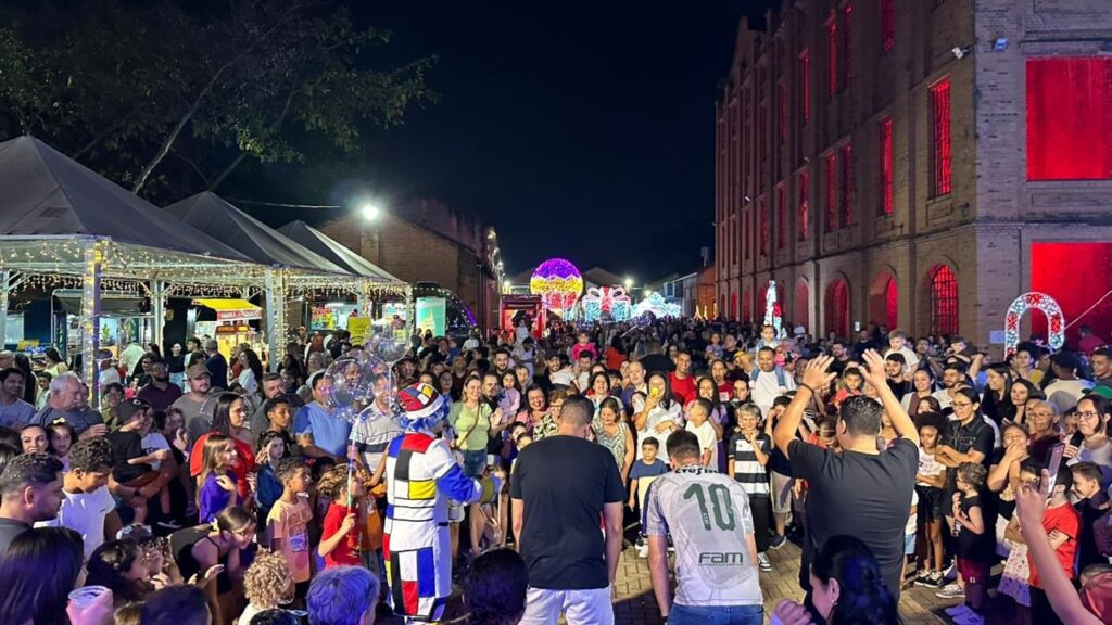 A imagem retrata uma vibrante cena noturna, possivelmente de um festival ou evento comemorativo. Uma multidão de pessoas, incluindo muitas crianças, está reunida em uma área externa, observando um espetáculo ou atração. À esquerda, barracas decoradas com luzes de Natal sugerem um mercado ou área de alimentação. As luzes cintilantes criam uma atmosfera festiva e acolhedora. Ao fundo, estruturas com iluminação decorativa, como grandes presentes e esferas coloridas, indicam a temática festiva do evento, possivelmente relacionada ao Natal ou a outra celebração. A edificação à direita, com janelas iluminadas em vermelho, adiciona um toque dramático e imponente à cena. A iluminação geral da imagem é noturna, com as luzes artificiais predominando. No centro da multidão, um artista vestido de palhaço, com um traje colorido e geométrico, parece interagir com o público, possivelmente animando as crianças. A presença de balões transparentes reforça o clima de celebração infantil. A atmosfera geral é de alegria, união e entretenimento, com pessoas de todas as idades compartilhando um momento de lazer e diversão sob a iluminação festiva.