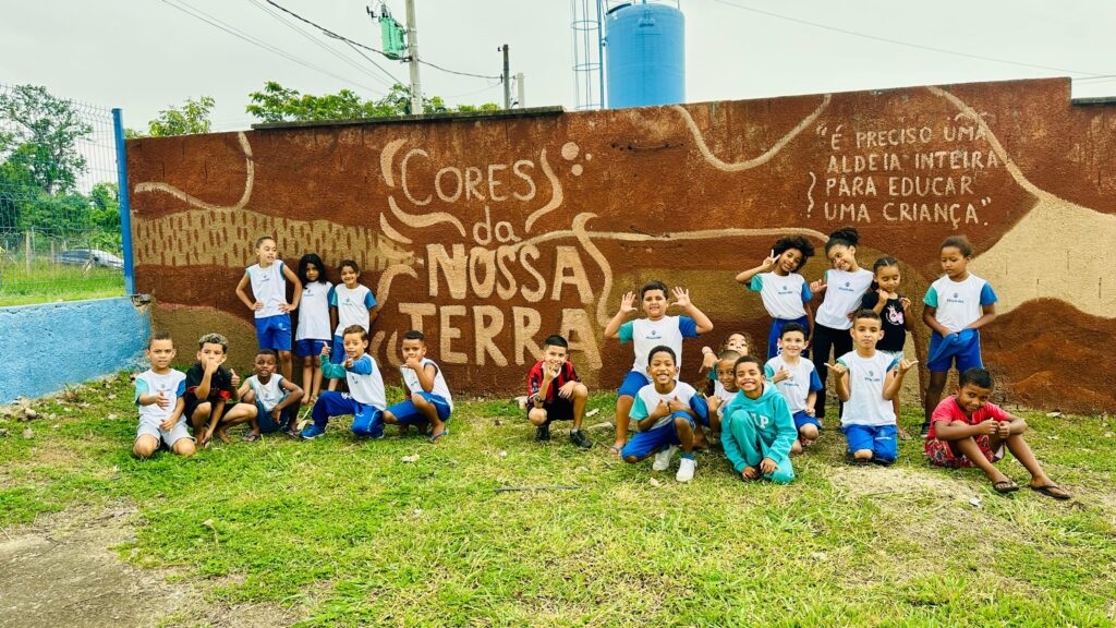 A imagem mostra um grupo de crianças em frente a um muro pintado com um mural. O mural tem a inscrição "CORES DA NOSSA TERRA" em letras grandes e estilizadas. À direita, há uma citação em português: "É preciso uma aldeia inteira para educar uma criança." As crianças, a maioria vestindo uniformes escolares brancos com detalhes azuis e shorts azuis, estão em diferentes posições: algumas em pé, outras agachadas ou sentadas na grama. Elas parecem estar em um ambiente externo, possivelmente uma escola ou área comunitária, com cercas e vegetação ao redor. O muro tem um fundo marrom texturizado, com elementos que lembram paisagens naturais ou texturas de terra. O céu está nublado.