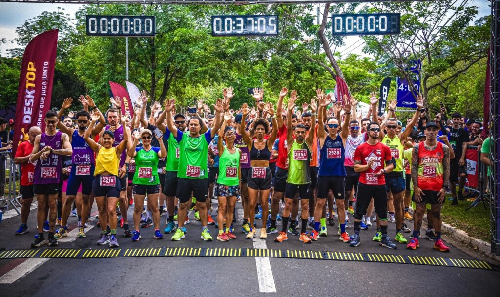 A imagem mostra um grupo de corredores reunidos na linha de largada de uma corrida. Eles estão posando para uma foto, com muitos deles com as mãos levantadas e sorrindo. Três cronômetros digitais são visíveis acima deles, mostrando tempos de 00:00:00, 00:37:05 e 00:00:00. Há bandeiras e banners ao redor da área de largada, com logotipos de patrocinadores como "DESKTOP" e "dontoclinic". Os corredores estão vestindo uma variedade de roupas de corrida coloridas, e muitos usam números de peito visíveis. A cena geral é vibrante e cheia de energia, capturando o entusiasmo antes do início de um evento esportivo.