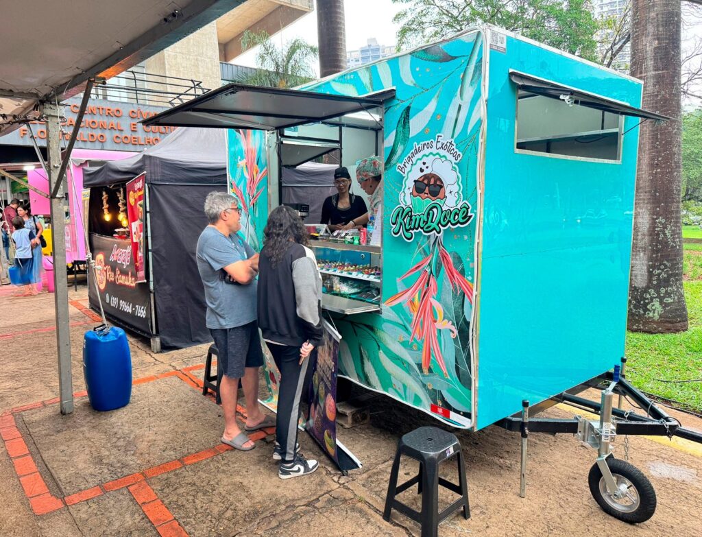 A imagem mostra uma feira de comida de rua com várias barracas. No centro da imagem, há uma barraca de comida vibrante e colorida, pintada de azul-turquesa com um design tropical de folhas verdes e flores cor-de-rosa. A barraca se chama "KimDoce" e anuncia "Brigadeiros Exóticos". Duas pessoas estão em pé em frente à barraca, olhando para os doces expostos. Uma delas, um homem com óculos cinza e camiseta cinza, parece estar conversando com a outra pessoa. A outra barraca, mais à esquerda, é rosa e tem um letreiro que diz "Açaí Ree Samuka" e um número de telefone. Há também uma barraca preta com luzes penduradas, possivelmente vendendo outros tipos de comida. O chão é pavimentado com tijolos e há algumas árvores e edifícios ao fundo, sugerindo um ambiente urbano. Um barril azul está próximo à barraca de açaí.