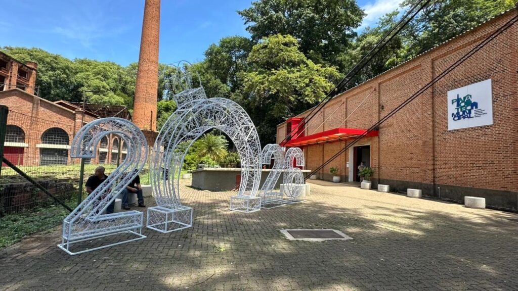 A imagem mostra uma instalação de arte externa com números grandes feitos de luzes brancas, formando o número "2026". Os números são esculturais e parecem ser feitos de uma malha metálica com luzes incorporadas, dando-lhes um brilho etéreo. Ao fundo, há um edifício de tijolos vermelhos com janelas arqueadas e uma chaminé alta de tijolos, sugerindo um local histórico ou industrial. Uma entrada com um toldo vermelho vibrante convida à entrada do edifício. A área em frente ao edifício é pavimentada com pedras e há vegetação verde exuberante ao redor, incluindo árvores altas que criam uma sombra parcial. No lado esquerdo da imagem, há uma cerca de arame e algumas pessoas sentadas perto da base de um dos números, possivelmente instalando ou admirando a obra. No edifício, há um grande sinal branco com letras azuis e verdes que diz "PIXA TECH MUNICIPAL". O céu está claro e azul, indicando um dia ensolarado.