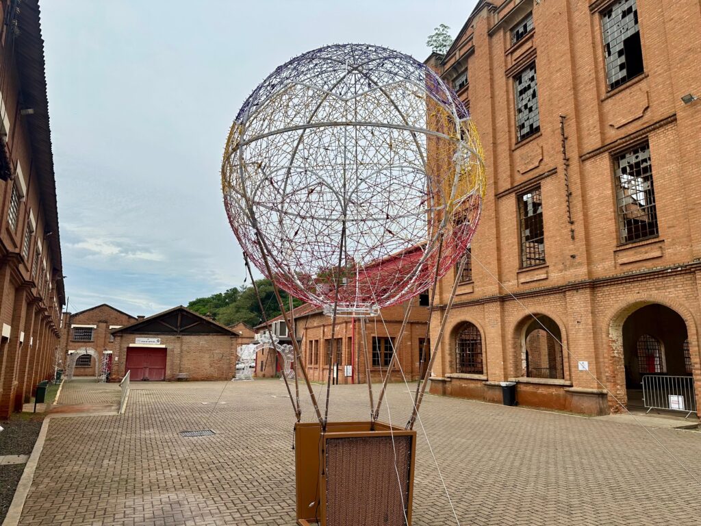 A imagem mostra uma instalação artística em forma de balão, feita de arame e luzes coloridas, em um pátio pavimentado com tijolos. O balão está suspenso por cabos e tem uma cesta de madeira na base. Ao fundo, erguem-se edifícios de tijolos aparentes, com janelas quebradas e um estilo arquitetônico industrial. A iluminação é suave, sugerindo um dia nublado ou o entardecer. A combinação do balão vibrante com a arquitetura rústica e um pouco decadente cria um contraste visual interessante.
