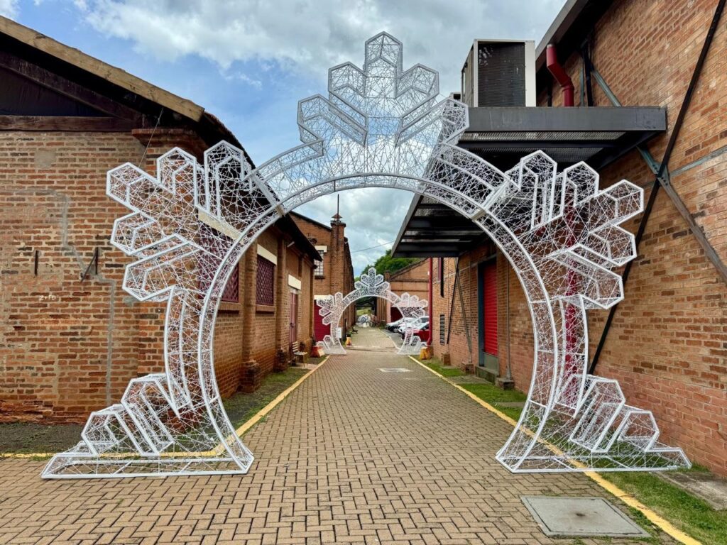 A imagem mostra um corredor ladeado por edifícios de tijolos vermelhos, com um céu nublado acima. No primeiro plano, há um grande arco decorativo branco feito de luzes, que se assemelha a um floco de neve estilizado. O arco é intrincadamente trabalhado com fios finos e parece ser uma instalação de arte ou decoração festiva. O corredor é pavimentado com tijolos e uma linha amarela marca a borda de um lado. Mais adiante no corredor, um arco menor semelhante pode ser visto, sugerindo que esta pode ser uma série de decorações. As paredes de tijolos dos edifícios são visíveis em ambos os lados, com janelas e portas vermelhas em alguns pontos. Há também um telhado inclinado de madeira em um dos edifícios à esquerda e unidades de ar condicionado montadas na parede de um edifício à direita. O ambiente geral é de um espaço ao ar livre, possivelmente em um centro cultural, parque ou área de eventos, decorado para uma ocasião especial.