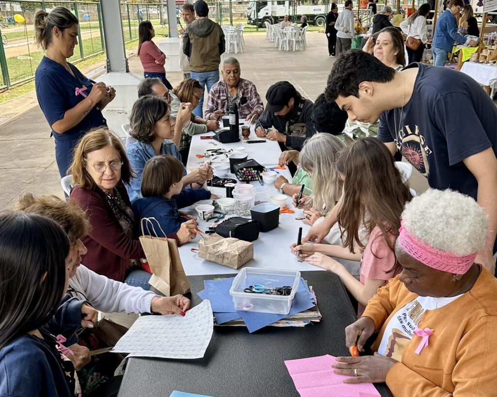 A imagem retrata um grupo diversificado de pessoas reunidas em torno de uma mesa, engajadas em uma atividade de artesanato ao ar livre. A cena, iluminada pela luz do dia, sugere um ambiente comunitário e colaborativo. No centro da imagem, a mesa está repleta de materiais para artesanato, como papéis coloridos, tesouras, canetas, fios e recipientes diversos. As mãos das pessoas estão ocupadas, cortando, desenhando e montando peças, demonstrando concentração e criatividade. Há uma variedade de idades e gêneros entre os participantes, desde crianças até idosos, todos compartilhando o mesmo espaço e interesse. Ao fundo, observa-se uma área externa com árvores, gramado e outras pessoas em atividades diversas, indicando um evento ou encontro maior. A atmosfera geral é de descontração e engajamento social, com um toque de aprendizado e expressão artística.