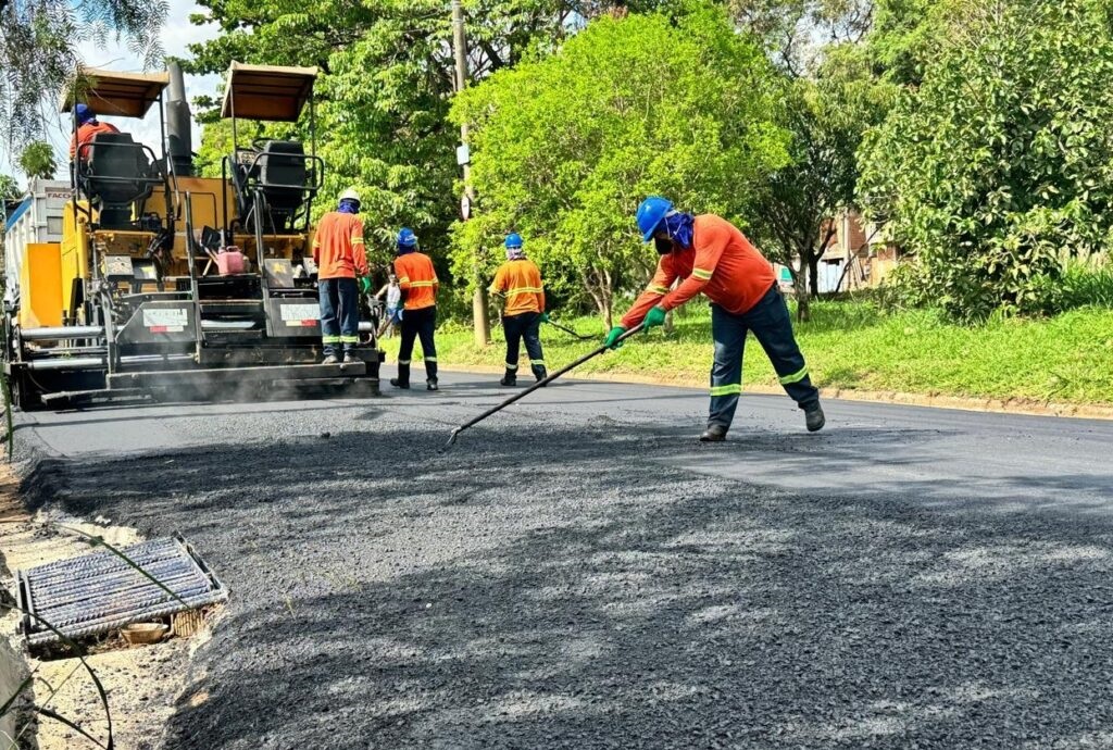 A imagem mostra uma equipe de trabalhadores realizando a pavimentação asfáltica de uma estrada. No centro da imagem, uma máquina pavimentadora está espalhando o asfalto quente, que emite vapor. Vários trabalhadores, vestidos com uniformes de segurança laranja e capacetes azuis, estão operando a máquina e guiando o asfalto com ferramentas. Um trabalhador em primeiro plano está usando um ancinho longo para nivelar o asfalto recém-colocado. Ao fundo, há uma área arborizada com árvores verdes e uma vegetação exuberante, indicando um ambiente externo e possivelmente rural ou suburbano. A cena retrata um processo de construção ou manutenção de infraestrutura viária.