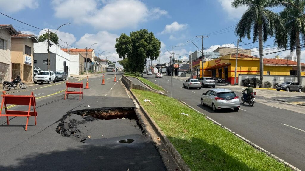 Na imagem, vê-se uma rua urbana onde um grande buraco se abriu em uma das faixas, formando uma cratera que compromete o asfalto. O local está isolado com barreiras vermelhas e cones laranja, garantindo a segurança dos motoristas e pedestres. O tráfego segue normalmente pela pista oposta, separada por um canteiro gramado. Ao redor, há prédios comerciais e residenciais, incluindo uma construção amarela e vermelha em destaque à direita. O céu parcialmente nublado completa o cenário, que evidencia um problema de infraestrutura e a necessidade de manutenção viária para evitar riscos à circulação.