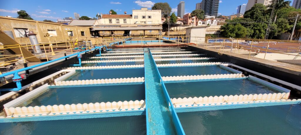A imagem mostra um sistema de tratamento de água, especificamente um conjunto de decantadores ou tanques de sedimentação. * A estrutura principal consiste em vários tanques longos e estreitos, dispostos em fileiras. * A água, de cor azul-esverdeada, preenche esses tanques. * As bordas superiores dos tanques são revestidas com um material branco com um padrão decorativo de "ondas" ou "dentes de serra". * Uma calha azul brilhante corre no centro da imagem, levando água para os tanques. * Ao fundo, há edifícios industriais e residenciais, com um céu azul claro e algumas nuvens. * A infraestrutura inclui tubulações azuis e amarelas, corrimãos e plataformas de acesso, indicando que se trata de uma instalação em funcionamento.