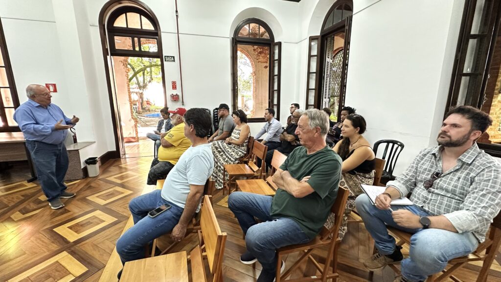 A imagem mostra um grupo de pessoas reunidas em uma sala com piso de madeira detalhado e janelas em arco. Um homem mais velho, vestindo uma camisa azul e jeans, está em pé e gesticulando, parecendo estar apresentando algo para o grupo. As outras pessoas estão sentadas em cadeiras de madeira, ouvindo atentamente. Alguns indivíduos estão mais próximos do foco, enquanto outros estão mais ao fundo, compondo a audiência. A luz entra pelas janelas, iluminando a cena e criando um ambiente acolhedor.