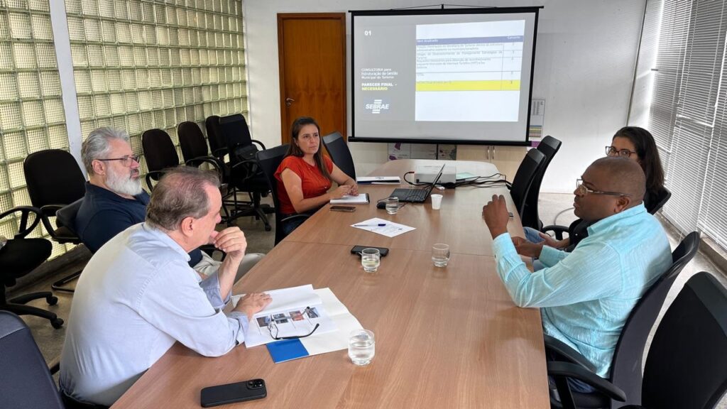 A imagem mostra um grupo de pessoas em uma sala de reuniões, sentadas ao redor de uma mesa de madeira. Há um projetor exibindo uma tela com texto e gráficos, possivelmente uma apresentação. Uma mulher de blusa laranja está sentada à esquerda, olhando para a tela. Um homem de camisa azul clara está à direita, gesticulando enquanto fala. Outros participantes estão visíveis, incluindo um homem com barba grisalha e óculos à esquerda e uma mulher com óculos atrás do homem de camisa azul. A sala parece ser um ambiente de escritório, com janelas de vidro e cadeiras de escritório. Há copos de água e papéis sobre a mesa, indicando uma reunião em andamento.
