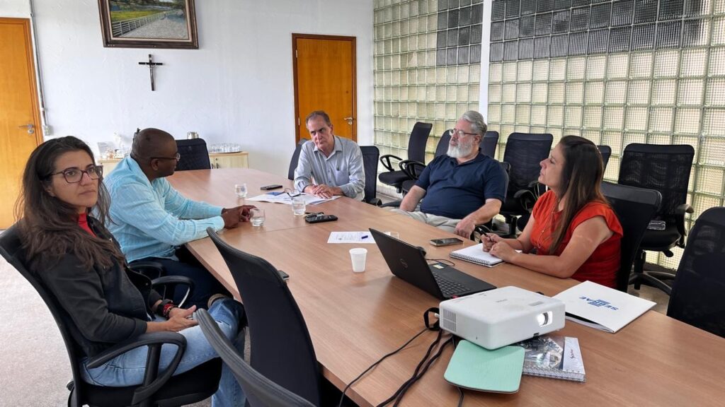 A imagem mostra uma sala de reuniões com cinco pessoas sentadas ao redor de uma mesa de madeira. No lado esquerdo da mesa, uma mulher com óculos e cabelos escuros está sentada, vestindo uma jaqueta preta sobre uma blusa vermelha e jeans. Ao lado dela, um homem com óculos e camisa azul clara conversa, olhando para a mesa. Na outra extremidade da mesa, um homem com camisa clara e cabelos curtos está sentado, olhando para a frente. Ao lado dele, um homem mais velho com barba grisalha e camisa azul escura está sentado. Na frente dele, uma mulher com cabelos castanhos e vestindo uma blusa laranja está sentada, olhando para a mesa. Na mesa, há copos de água, papéis, cadernos, um laptop aberto, um celular e um projetor. Ao fundo, há uma porta de madeira e uma parede com blocos de vidro translúcido. Acima da porta, há um quadro com uma paisagem e uma cruz pendurada na parede.