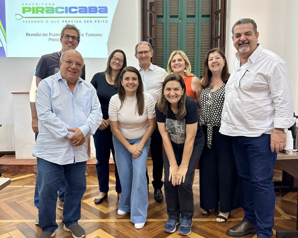 A imagem mostra um grupo de nove pessoas em pé, em um ambiente interno, possivelmente uma sala de reuniões, com um projetor ao fundo exibindo um slide com o logo da prefeitura de Piracicaba. As pessoas estão sorrindo e olhando para a câmera, sugerindo um momento de confraternização ou uma foto oficial. Os indivíduos vestem roupas casuais e formais, com predominância de camisas de botão e calças jeans ou sociais. A iluminação é suave, vinda de cima e da frente, destacando os rostos e as expressões de alegria do grupo. O piso de madeira com um padrão em espinha confere um toque clássico ao ambiente.