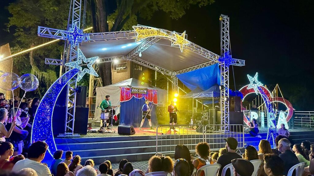 A imagem mostra uma cena noturna de um palco ao ar livre, decorado com luzes festivas. No centro, três artistas se apresentam em um palco elevado com uma cortina vermelha e azul ao fundo, onde se lê "RISO SHOW". Um deles toca guitarra e canta, outro toca um instrumento de sopro (possivelmente um trombone), e o terceiro parece estar falando ou cantando. O palco é ladeado por estruturas metálicas com decorações luminosas em forma de estrelas azuis e amarelas, e arcos com luzes azuis e vermelhas. Uma multidão de pessoas assiste à apresentação sentada em cadeiras e em pé, segurando balões transparentes. A iluminação do palco é amarelada, criando um ambiente vibrante e animado. Árvores e construções ao fundo, juntamente com as luzes, sugerem um evento comunitário ou festival.