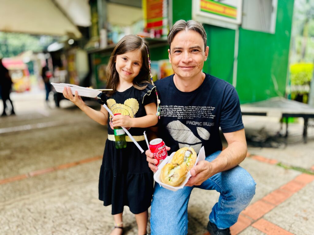 A imagem mostra um homem e uma menina em pé, em frente a barracas de comida. O homem, ajoelhado, segura um sanduíche e uma lata de Coca-Cola. A menina, ao lado dele, segura um prato com comida e uma garrafa de bebida. Ambos sorriem para a câmera. O homem veste uma camiseta preta com uma estampa de folhas e a escrita "PACIFIC BLUE", e calça jeans. A menina usa um vestido preto com detalhes dourados, que parecem ser de um urso. O fundo da imagem é desfocado, mas é possível ver outras barracas de comida e pessoas. A iluminação sugere que é dia.