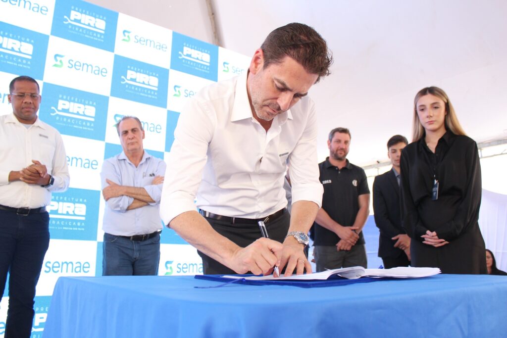 A imagem mostra um evento oficial onde um homem de camisa branca assina um documento em uma mesa coberta com uma toalha azul. Ele está no centro da imagem, curvado sobre o documento, com uma caneta na mão direita. Ao seu redor, várias pessoas estão em pé, observando o ato. No fundo, um painel com o logo da "Prefeitura de Piracicaba" e "Semae" está visível, indicando que o evento está relacionado a essas instituições. À esquerda do homem que assina, um homem de óculos e camisa branca está com as mãos juntas. Ao lado dele, um homem mais velho com camisa azul clara e braços cruzados observa atentamente. À direita do homem que assina, um homem de camiseta preta com o número "1913" e um jovem de terno observam. Uma mulher jovem com uma camisa preta está mais à direita, também observando. O ambiente parece ser ao ar livre, sob uma estrutura de cobertura.