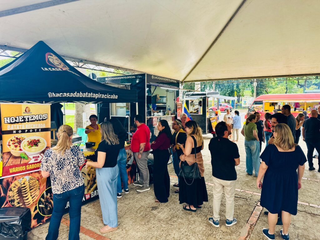 A imagem mostra um evento ao ar livre, possivelmente uma feira de rua ou festival gastronômico, sob uma grande tenda branca. Várias barracas de comida estão montadas, com destaque para uma tenda preta com o logo "LA CASA DO ESPETO" e o nome "@acasadoespeto.oficial" visível. Ao lado, há um banner amarelo vibrante anunciando "HOJE TEMOS! BATATA RECHEADA NO POTE", com imagens apetitosas de batatas recheadas e batatas fritas. Uma fila de pessoas se forma em frente à barraca, indicando a popularidade do local. Várias pessoas estão em pé, conversando e aguardando seus pedidos. Ao fundo, outros food trucks e barracas podem ser vistos, juntamente com algumas árvores e vegetação, sugerindo um ambiente de parque ou área verde. A atmosfera parece animada e social, com pessoas desfrutando da comida e da companhia umas das outras.