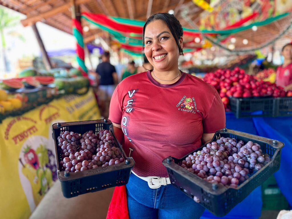 Uma mulher sorri para a câmera enquanto segura duas caixas de uvas roxas em um mercado de frutas. Ela está usando uma camiseta vermelha com um logo no peito que diz "COMÉRCIO DE FRUTAS FINOTTI". Atrás dela, há uma variedade de outras frutas, incluindo melancias e maçãs. As frutas estão dispostas em bancas sob uma cobertura de madeira, e há decorações verdes e vermelhas penduradas no teto. O ambiente parece ser um mercado ao ar livre, com outras pessoas ao fundo.