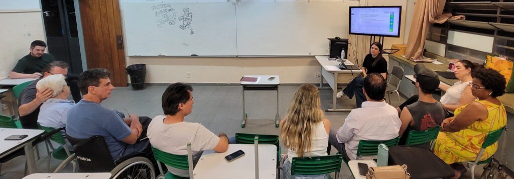 A imagem mostra uma sala de aula com várias pessoas sentadas em cadeiras, voltadas para uma tela de projeção. Uma mulher está sentada perto da tela, aparentemente apresentando algo. Há também um quadro branco com um desenho de um jogador de futebol e a frase "ELETIVA FUTEBOL EM JOGO!". Algumas mesas estão dispostas na sala, com laptops e celulares sobre elas. Uma pessoa em cadeira de rodas está visível em primeiro plano.