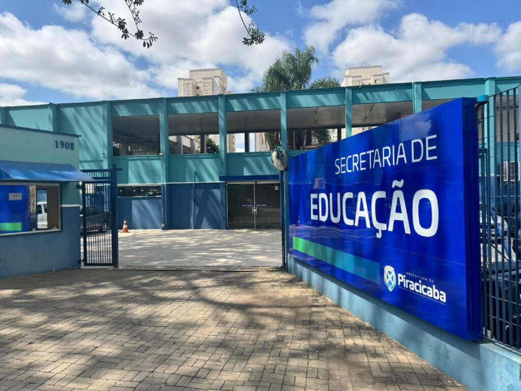 A imagem mostra a fachada de um prédio, identificado como a "Secretaria de Educação" da Prefeitura de Piracicaba. O edifício é pintado em tons de azul e verde-água, com uma arquitetura moderna. Em primeiro plano, um grande letreiro azul com letras brancas exibe o nome da secretaria e o brasão da prefeitura. À esquerda, um portão gradeado dá acesso ao interior do prédio, onde se vê uma pequena guarita. O número "1902" está visível em uma das paredes. O céu, com nuvens brancas, sugere um dia claro. O chão em frente ao prédio é pavimentado com blocos de concreto em tom ocre.