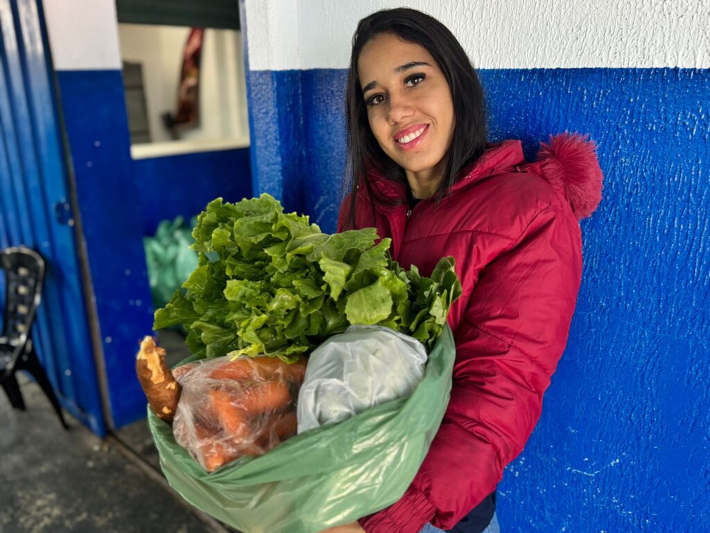 A foto mostra uma pessoa vestindo um casaco vermelho de inverno, em pé diante de uma parede azul e branca, segurando uma sacola plástica verde cheia de mantimentos. Dentro da sacola é possível ver verduras frescas, como alface, além de cenouras, pão e outros itens embalados.