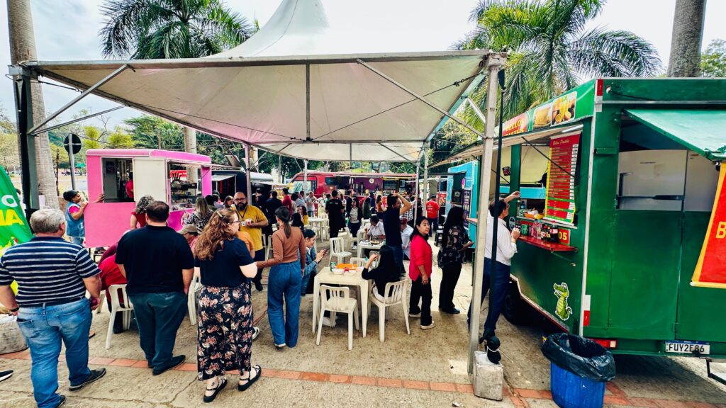 A imagem mostra um evento comunitário animado em uma área externa, com vários food trucks coloridos alinhados sob uma grande tenda. As pessoas circulam entre os veículos, algumas pedindo comida, outras sentadas em mesas e cadeiras plásticas brancas para se alimentar. O ambiente é cercado por palmeiras, reforçando a atmosfera de clima quente e descontraído. Os caminhões, em tons de verde, rosa, vermelho e azul, dão um ar vibrante ao espaço, transmitindo a ideia de um festival gastronômico urbano marcado pela interação social e pela diversidade de sabores.