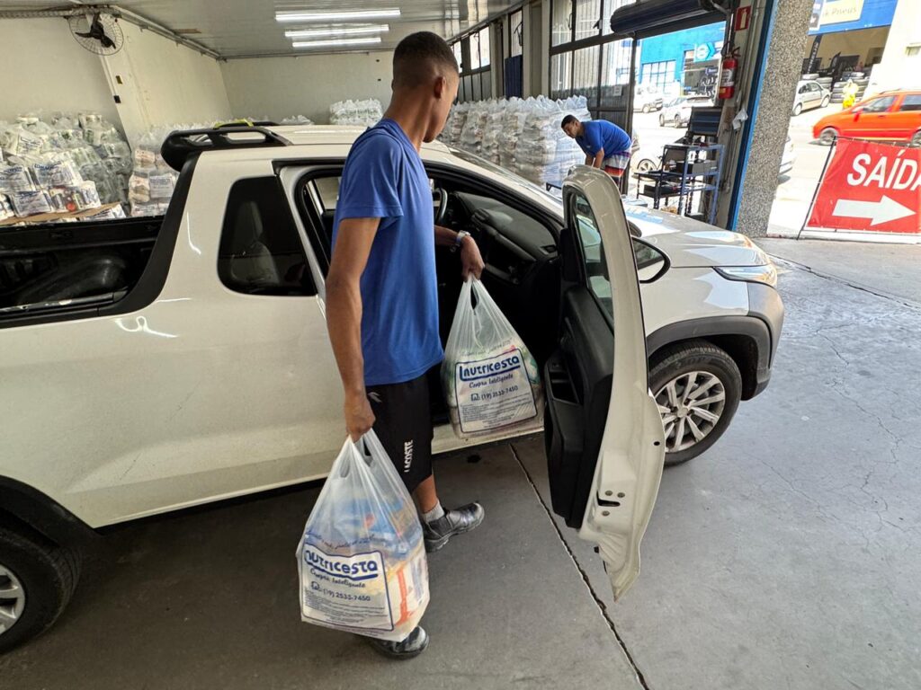 A imagem mostra duas pessoas carregando sacolas de alimentos embalados em uma caminhonete branca, em um espaço que parece ser um centro de distribuição ou armazém. Em primeiro plano, um homem coloca duas grandes sacolas identificadas como “Nutricesta Compra Inteligente” dentro do veículo, enquanto ao fundo outro trabalhador organiza mais pacotes próximos a uma pilha de mantimentos.