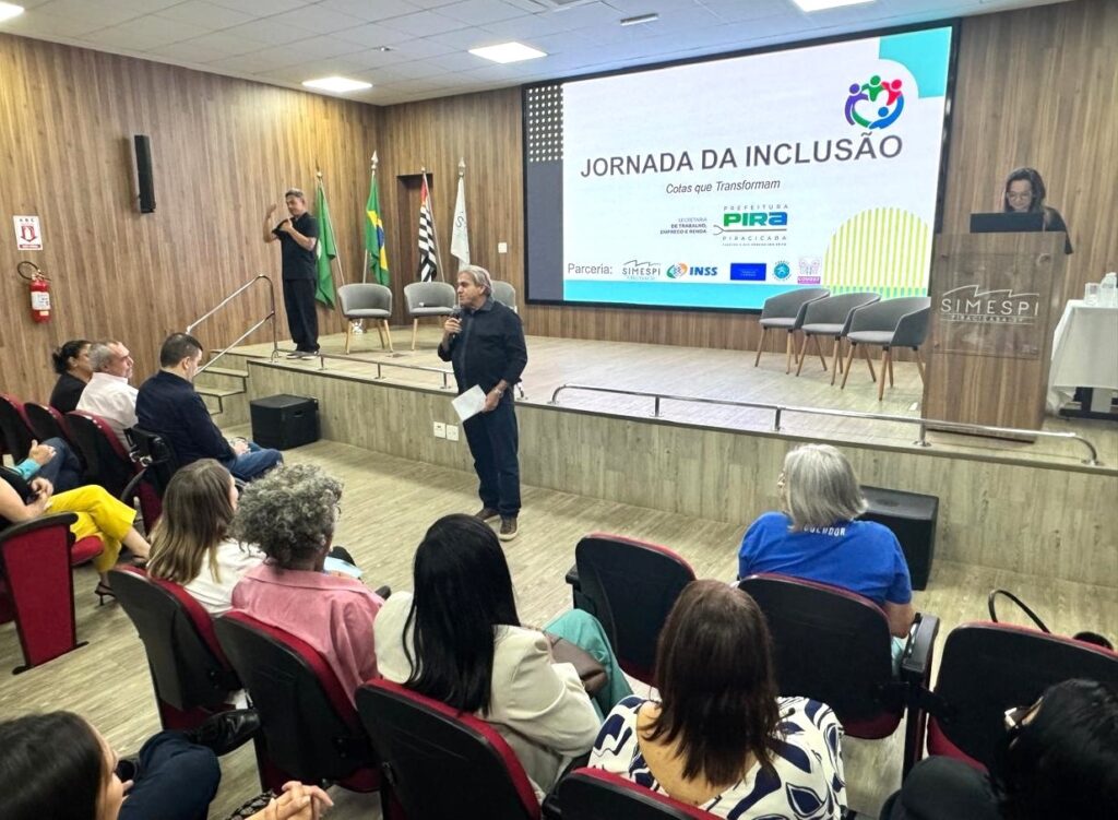 A imagem mostra um seminário em auditório, com um palestrante falando ao público enquanto segura um microfone e um papel. Ao fundo, um grande telão exibe o título do evento: “Jornada da Inclusão – Cotas que Transformam”, acompanhado dos logotipos de instituições parceiras como SIMESPI, INSS, Prefeitura de Piracicaba, Secretaria Municipal de Desenvolvimento Econômico, Trabalho e Turismo, Ministério do Trabalho e Emprego e Superintendência Regional do Trabalho em São Paulo. No palco, também há uma pessoa utilizando a Língua de Sinais, reforçando o caráter inclusivo da iniciativa. O público está acomodado em poltronas vermelhas, em um espaço moderno com painéis de madeira. A cena transmite a ideia de um encontro formal voltado à promoção da inclusão e da diversidade no mercado de trabalho.