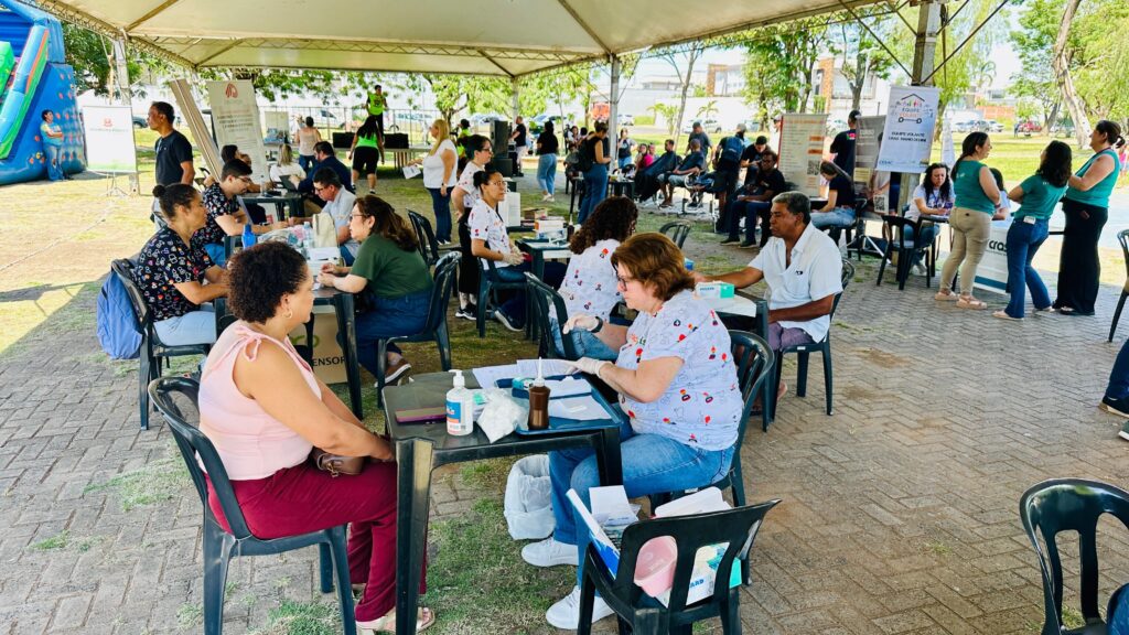 Na imagem, vê-se uma feira comunitária de saúde realizada em um espaço aberto, sob uma grande tenda. Diversas mesas estão organizadas com profissionais de saúde atendendo moradores, usando uniformes e luvas, em atividades como consultas, triagens e orientações.