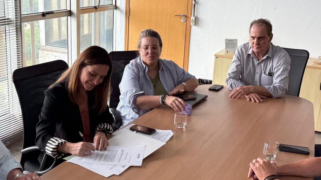 À esquerda, uma mulher com cabelo castanho e um blazer preto está assinando um documento. Ela está sorrindo levemente enquanto se concentra na tarefa. No centro, uma mulher com óculos e cabelo loiro está sentada, olhando para a frente. Ela usa uma camisa listrada azul e branca sobre uma blusa verde e está usando várias pulseiras em seu pulso. À direita, um homem com cabelo castanho e uma camisa cinza abotoada está sentado com os braços cruzados sobre a mesa. Ele parece estar observando a mulher que está assinando o documento. Há vários telefones celulares e um copo de água na mesa. As janelas ao fundo mostram uma vista de árvores e edifícios, sugerindo que a sala de reuniões está em um andar superior.