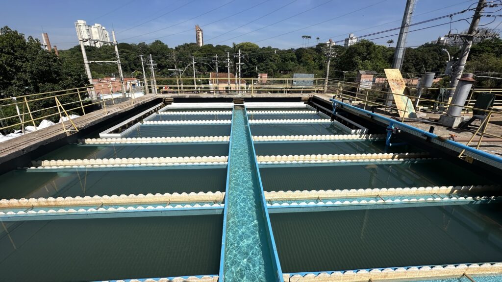 A imagem mostra uma grande estação de tratamento de água ao ar livre, com várias tanques retangulares paralelos preenchidos com água esverdeada e turva. Entre os tanques, há passarelas de concreto e um canal central pintado de azul, por onde a água parece estar fluindo. As bordas dos tanques têm um acabamento branco com ondulações. A foto foi tirada de um ângulo alto, permitindo uma visão ampla da instalação. Ao fundo, é possível ver árvores verdes, postes de energia elétrica com fios, e alguns edifícios, incluindo um prédio residencial alto e moderno e uma estrutura industrial menor com chaminés. O céu está claro e azul, indicando um dia ensolarado, provavelmente durante a manhã ou início da tarde. Não há pessoas na cena, e a imagem transmite a ideia de um processo industrial de tratamento de água em funcionamento.