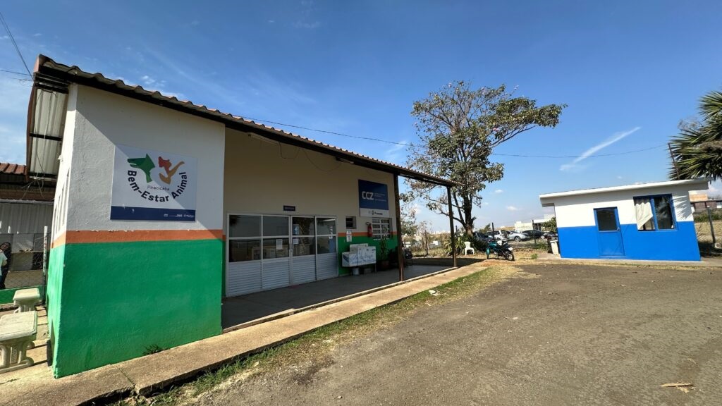A imagem mostra a fachada de um edifício com a inscrição "Bem-Estar Animal" e um logotipo com silhuetas de animais. O edifício é pintado de branco com listras verdes e laranjas na parte inferior. Há uma varanda com colunas de sustentação e telhado. Ao lado, há uma pequena construção pintada de branco e azul, com uma porta e uma janela. O céu está azul e claro, com algumas nuvens dispersas. Há árvores e vegetação ao redor, e alguns veículos estacionados ao fundo.