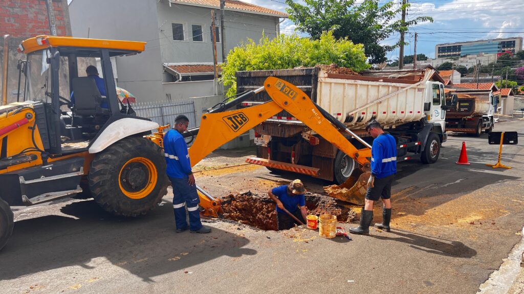 A imagem mostra uma cena de trabalho em uma rua, onde uma escavadeira amarela da marca JCB está sendo utilizada para cavar um buraco no asfalto. Ao lado da escavadeira, um caminhão basculante branco está posicionado, com a caçamba cheia de terra, indicando que o material escavado está sendo transportado. Três trabalhadores, vestidos com uniformes azuis e calças de trabalho com faixas refletivas, estão envolvidos na operação. Um deles está operando a escavadeira, visível através da cabine. Outro trabalhador está em pé ao lado da máquina, observando o trabalho. O terceiro trabalhador está dentro do buraco cavado, utilizando uma pá para remover terra, enquanto um balde amarelo e outros materiais de construção estão próximos. Ao fundo, observam-se casas com telhados laranjas e árvores frondosas, além de um edifício moderno com várias janelas envidraçadas, sugerindo que a obra está ocorrendo em uma área urbana. Há também cones de trânsito e sinalização amarela na rua, indicando que a área de trabalho está isolada para segurança. O céu está parcialmente nublado, com um clima que parece ser de dia.