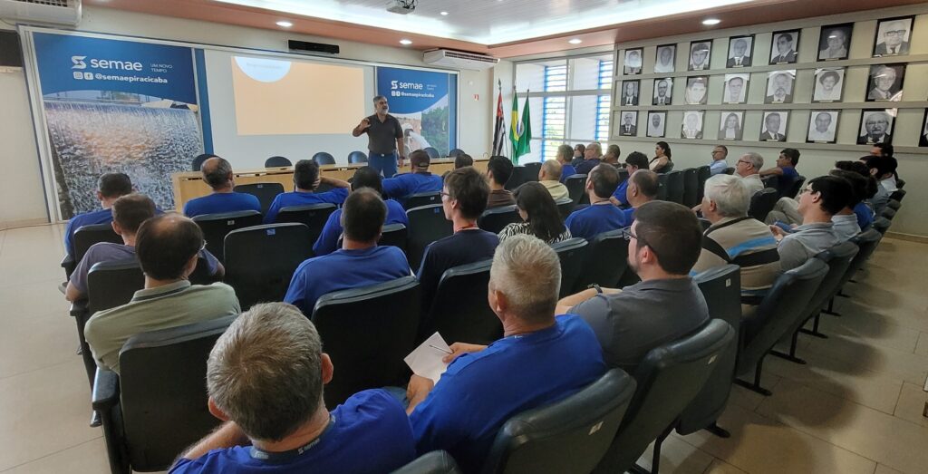 A imagem mostra uma sala de conferências onde um homem está em pé em frente a uma plateia sentada. O homem em pé, que parece ser o orador, está vestindo uma camisa escura e calças azuis e está gesticulando com as mãos enquanto fala. Ele está posicionado em frente a um pôster com o logotipo "semae" e a frase "UM NOVO TEMPO". A plateia é composta principalmente por homens, a maioria vestindo camisetas azuis. Eles estão sentados em cadeiras em fileiras, prestando atenção ao orador. Ao fundo, há uma parede adornada com uma série de retratos emoldurados, possivelmente de figuras importantes ou ex-funcionários. Bandeiras do Brasil e do estado de São Paulo podem ser vistas ao lado do orador. A iluminação da sala é artificial, vinda de luminárias no teto.