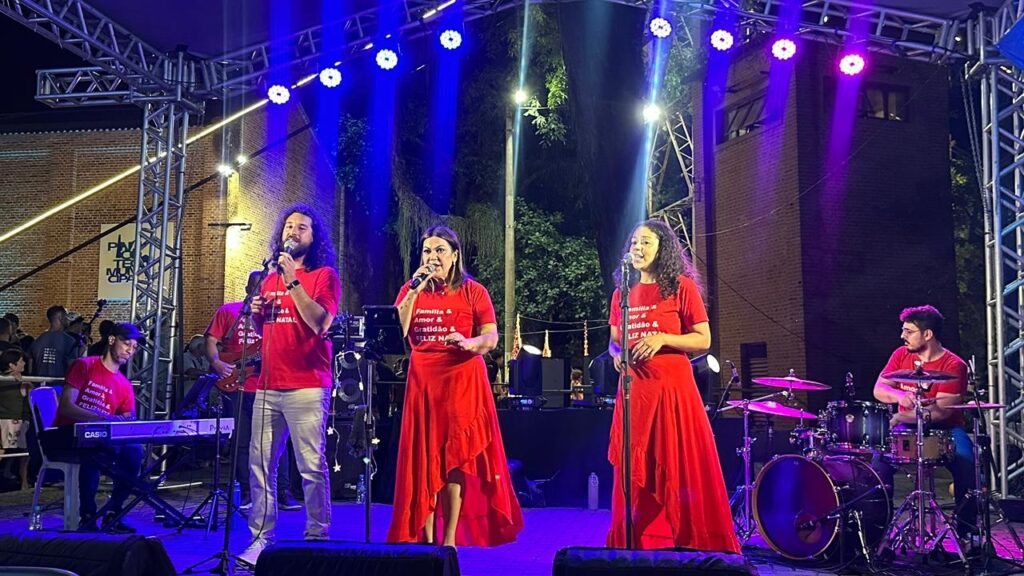 A imagem mostra uma banda se apresentando em um palco ao ar livre à noite. Há uma estrutura metálica com luzes azuis e roxas iluminando o palco. No centro, três cantores (dois mulheres e um homem) estão usando camisetas vermelhas com a inscrição "Família & Amor & Gratidão & Feliz Natal". Eles estão cantando em microfones. À esquerda, um homem toca teclado, e à direita, um baterista toca bateria. Ao fundo, outras pessoas podem ser vistas, e há uma parede de tijolos e árvores. A atmosfera parece ser festiva, possivelmente um evento de Natal.