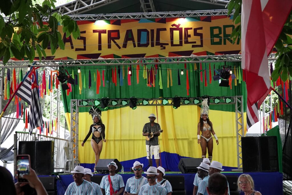 A imagem mostra uma cena vibrante de um palco montado ao ar livre, possivelmente para um festival ou celebração. No centro do palco, há um músico tocando um instrumento de cordas, que parece ser um ukulele. Ao lado dele, duas dançarinas em trajes de carnaval deslumbrantes, com penas e pedrarias, posam com confiança. O fundo do palco é decorado com cortinas verdes e amarelas, remetendo às cores da bandeira do Brasil. Penduradas acima do palco, há uma faixa com a inscrição "AS TRADIÇÕES BR" em letras grandes e coloridas, e uma série de fitas coloridas em tons vibrantes como laranja, rosa, verde e azul. À esquerda do palco, uma bandeira com listras pretas e brancas e um detalhe vermelho se destaca, possivelmente representando um estado brasileiro ou uma bandeira histórica. À direita, uma bandeira com listras vermelhas e brancas também está visível.