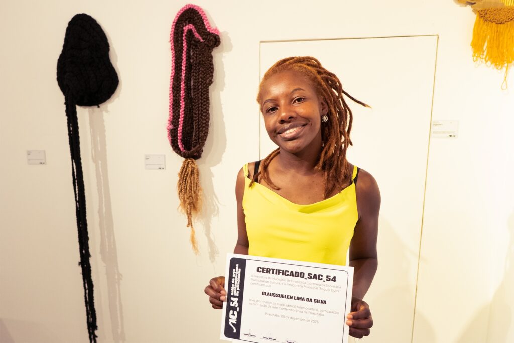 A mulher está em pé em frente a uma parede branca, sorrindo para a câmera. Ela está usando um top amarelo vibrante e tem dreadlocks cor de gengibre. Ela está segurando um certificado na mão direita. Atrás dela, penduradas na parede, há duas peças de arte têxtil em forma de cápsula. A peça à esquerda é preta e tem um longo pingente preto. A peça à direita é marrom com detalhes em rosa e tem um pingente de franjas cor de caramelo. Há também uma peça de arte têxtil amarela pendurada na parede à direita, parcialmente visível. A iluminação é suave e cria sombras suaves nas obras de arte.