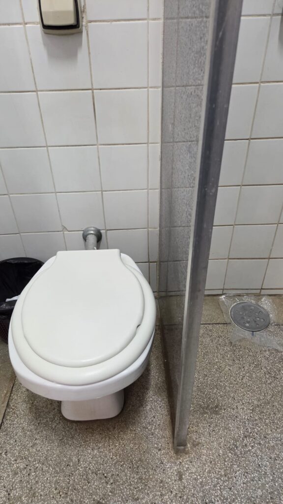 A imagem mostra um banheiro com azulejos brancos nas paredes e um piso de granilite. Há um vaso sanitário branco com a tampa fechada, posicionado à esquerda. Ao lado do vaso, há uma lixeira preta. Uma divisória de vidro com estrutura metálica divide o espaço, e à direita do vaso, no piso, há um ralo com uma tampa metálica circular. A iluminação parece ser artificial, com uma luz embutida visível na parte superior esquerda da imagem.